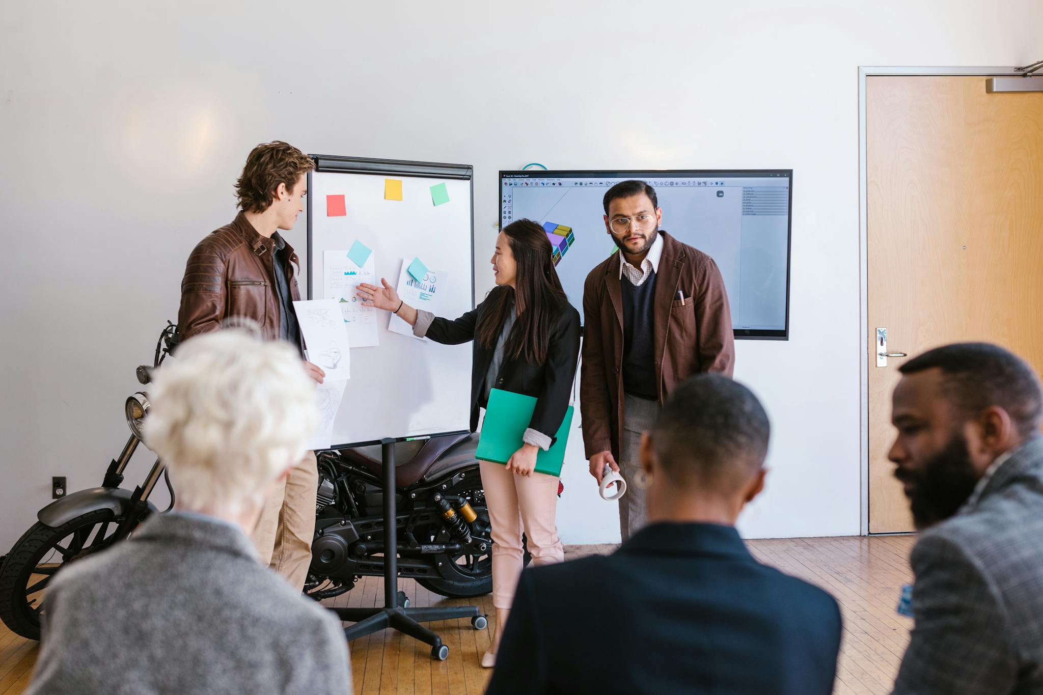 A multicultural team collaborates in an office, presenting ideas using a whiteboard and digital screen.