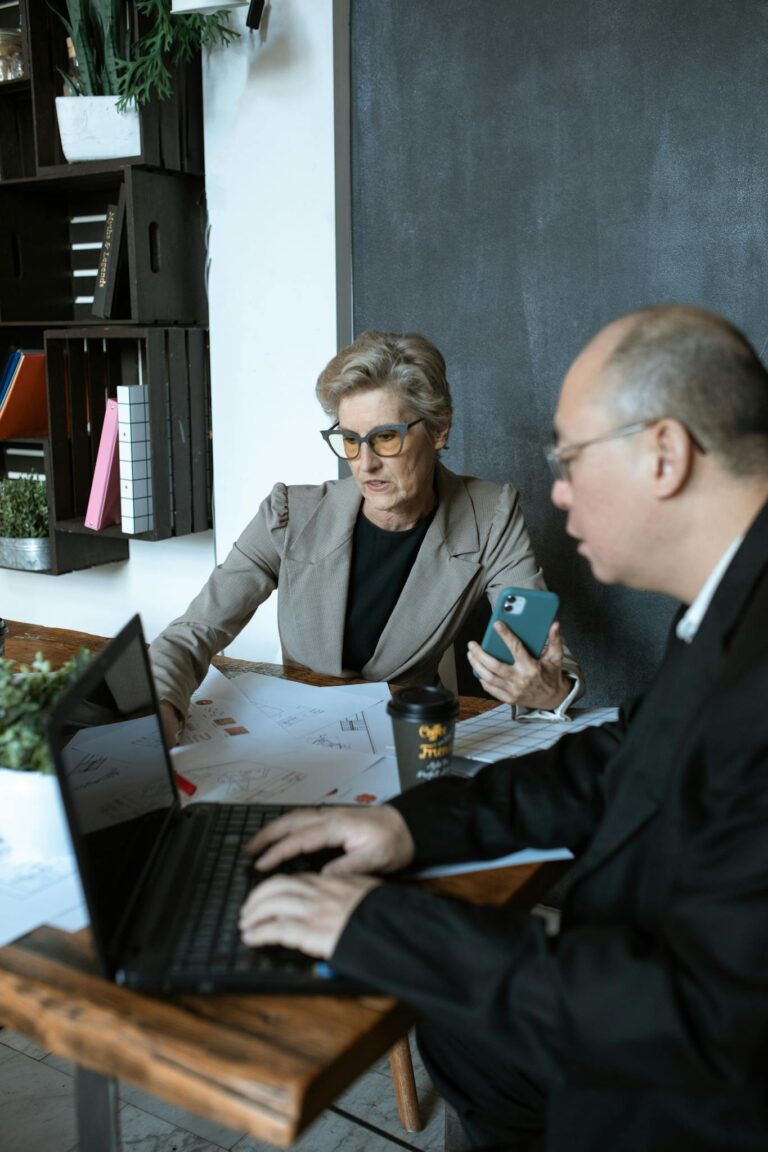 Two senior colleagues discussing a project in a modern office setting, using laptops and mobile devices.